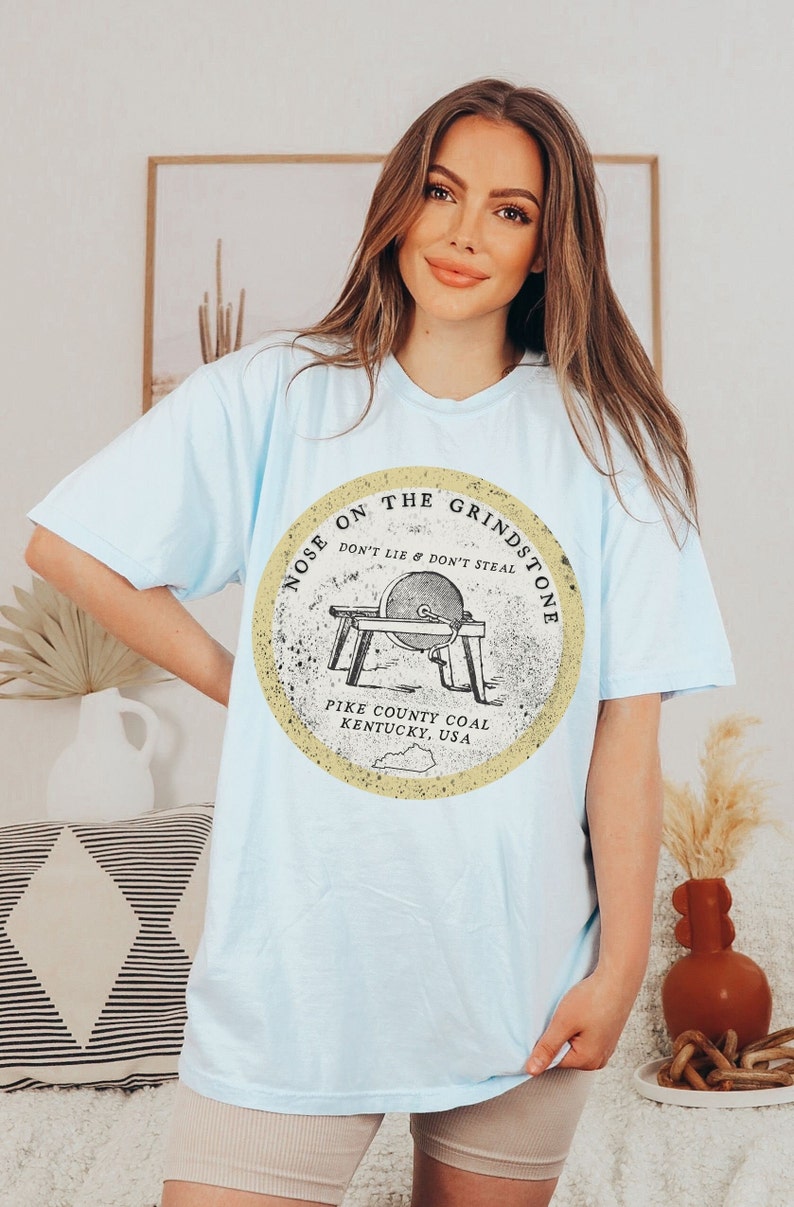 May include: Light blue t-shirt with a vintage-style graphic of a grindstone with the text "Nose on the Grindstone Don't Lie & Don't Steal Pike County Coal Kentucky, USA."
