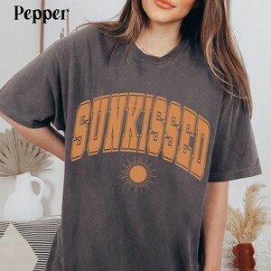 May include: A dark gray t-shirt with the word "SUNKISSED" printed in orange on the front. The word is in a bold, blocky font and is arched over a small orange sun graphic.