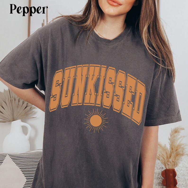 May include: A dark gray t-shirt with the word "SUNKISSED" printed in orange on the front. The word is in a bold, blocky font and is arched over a small orange sun graphic.