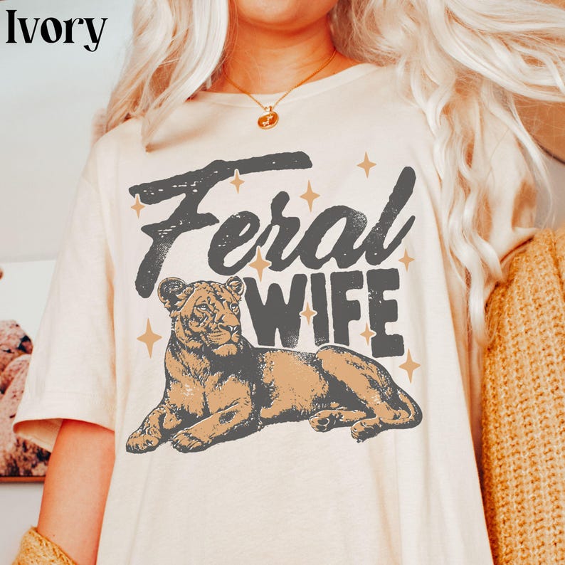 May include: A white t-shirt with the text "Feral Wife" in black lettering and a brown and black lion illustration. The shirt has a distressed vintage look.