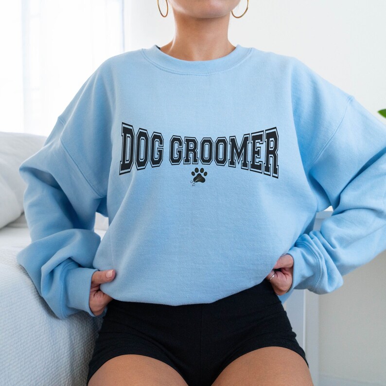 May include: A light blue sweatshirt with the text "DOG GROOMER" in black lettering with a paw print icon below. The sweatshirt is being worn by a person.