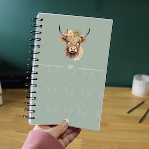 Puede incluir: Un cuaderno de espiral con una cubierta verde claro que presenta una ilustración de acuarela de una vaca de las Tierras Altas con una corona floral. El cuaderno tiene un diseño de corazón y un patrón floral en la mitad inferior.