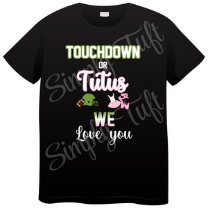 Op de afbeelding: Zwart T-shirt met de tekst "Touchdown or Tutus We Love You" in groen en roze. Een voetbalhelm en een paar roze balletten zijn afgebeeld.