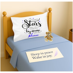 Peut inclure: Une taie d'oreiller blanche avec le texte "He counts the Stars and calls them all by name" et le nom "Michael" en bleu. Un petit ours en peluche est posé sur un lit bleu avec une note "Sleep in peace Wake in joy."