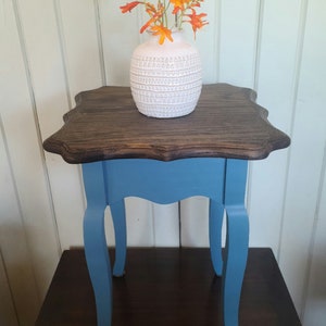 Puede incluir: Una pequeña mesa auxiliar azul con una tapa festoneada y tres patas. La mesa tiene una tapa de madera oscura y un jarrón de cerámica blanca con flores naranjas en la parte superior.