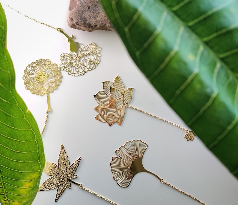 May include: Set of five gold metal bookmarks featuring different floral and leaf designs. The bookmarks are attached to chains and have a delicate, intricate look.