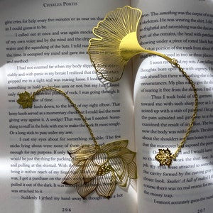 May include: Two gold metal bookmarks shaped like a ginkgo leaf and a lotus flower. The bookmarks are on an open book with text about a skeleton and a shirt. The text on the book reads "True Grit" and "Charles Portis".