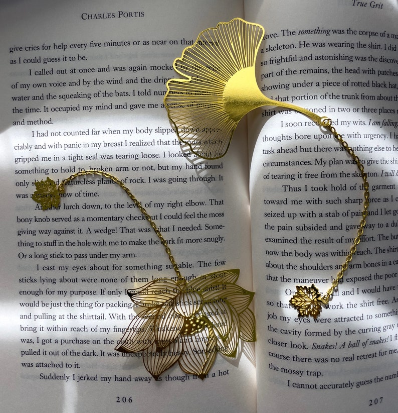 May include: Two gold metal bookmarks shaped like a ginkgo leaf and a lotus flower. The bookmarks are on an open book with text about a skeleton and a shirt. The text on the book reads "True Grit" and "Charles Portis".