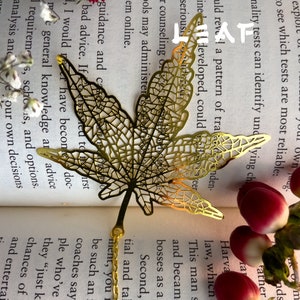 May include: A gold-colored metal bookmark shaped like a maple leaf. The bookmark is laying on a book with a white background and red flowers.