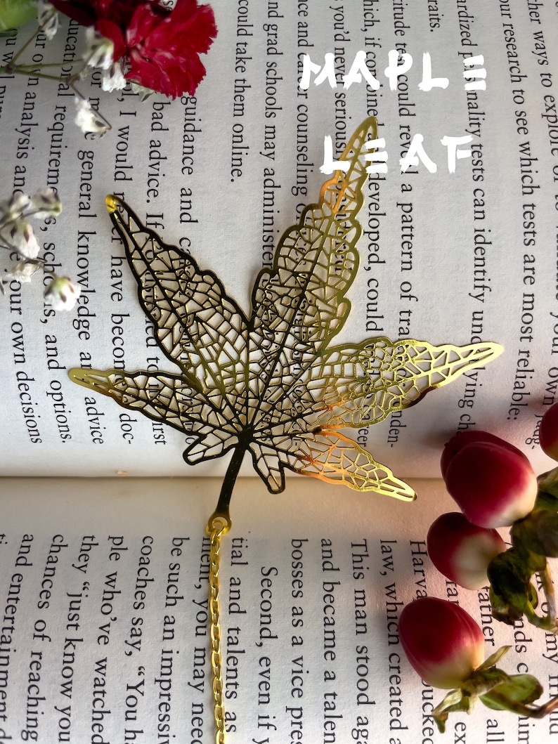 May include: A gold-colored metal bookmark shaped like a maple leaf. The bookmark is laying on a book with a white background and red flowers.