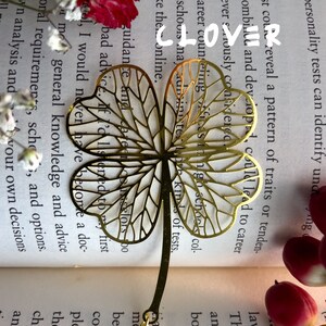 May include: A gold-colored four-leaf clover bookmark with a delicate, intricate design. The bookmark is made of metal and has a chain attached to the top.