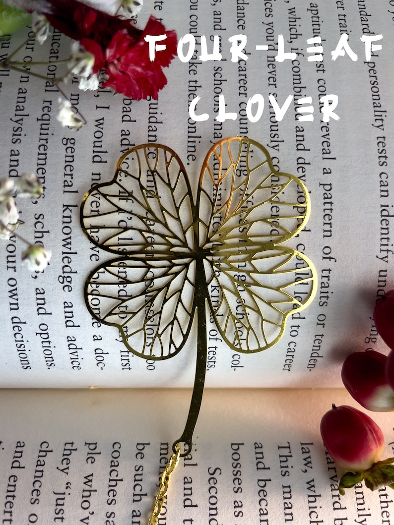 May include: A gold-colored four-leaf clover bookmark with a delicate, intricate design. The bookmark is made of metal and has a chain attached to the top.