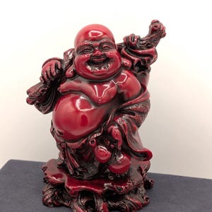 May include: A glossy, red resin Laughing Buddha statue. The figure is depicted with a large belly, smiling face, and is holding a staff and a bag. The statue is on a decorative base.