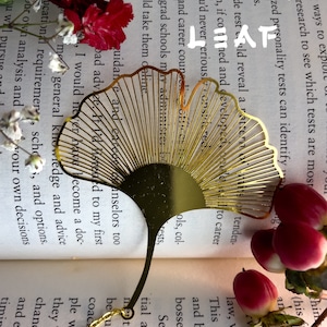 May include: A gold-colored metal bookmark shaped like a ginkgo leaf. The bookmark is laying open on a book with the text "GINKGO LEAF" visible. The bookmark is surrounded by pink flowers.