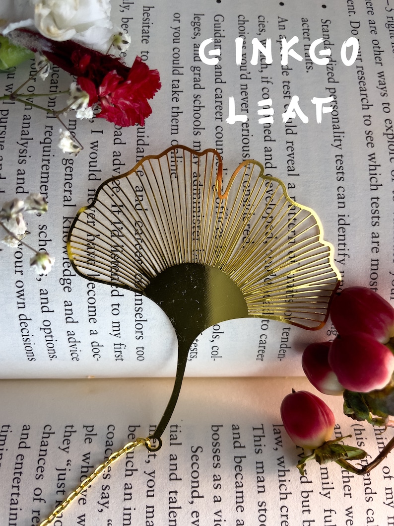 May include: A gold-colored metal bookmark shaped like a ginkgo leaf. The bookmark is laying open on a book with the text "GINKGO LEAF" visible. The bookmark is surrounded by pink flowers.