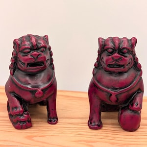 May include: Two maroon-colored Foo Dog statues. The statues are in a seated position with detailed facial features and a ball under one paw. The statues are on a wooden surface.