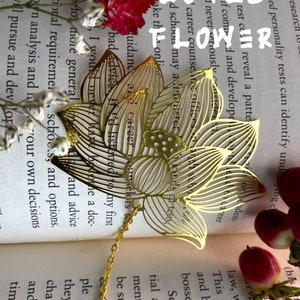 May include: A gold metal lotus flower bookmark with a chain attached. The bookmark is laying open on a book with white flowers and a red flower.