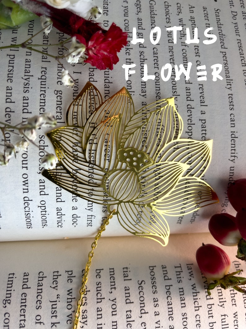 May include: A gold metal lotus flower bookmark with a chain attached. The bookmark is laying open on a book with white flowers and a red flower.