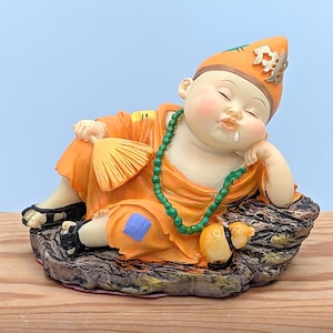 May include: A figurine of a sleeping baby monk in orange robes and a matching hat, resting on a rock-like base. The monk wears a green beaded necklace and black sandals. A small fan rests in the monk's hand. The figurine has a serene expression.
