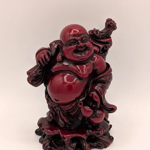May include: A glossy, red resin Buddha statue. The figure is depicted smiling, holding a staff, and standing on a detailed, textured base. The statue has a smooth, reflective surface and is a decorative item.