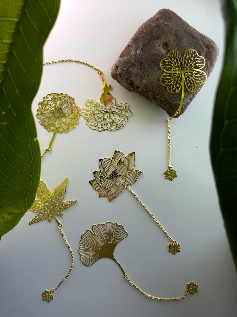 May include: A set of six gold-toned metal bookmarks featuring various floral and leaf designs. The bookmarks are attached to chains with small decorative charms at the ends.