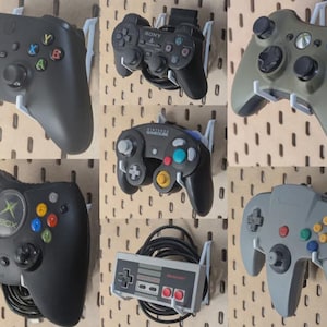 May include: A collection of seven video game controllers mounted on white plastic wall hooks. The controllers include various models from Xbox, Sony PlayStation, Nintendo GameCube, Nintendo NES, and Nintendo 64, featuring diverse button layouts.