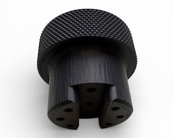 May include: A black cylindrical knob featuring a textured, diamond-patterned knurled top surface. The base of the knob has a recessed center section and includes six small circular holes arranged symmetrically around the bottom edge.