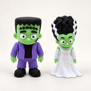 May include: Two stylized figurines featuring a green-skinned character with a black flat-top haircut, purple suit, and black shoes, standing next to a green-skinned character with a tall black and white beehive hairstyle wearing a white dress.
