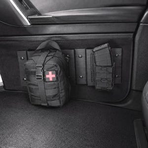 May include: A black modular storage panel mounted to a vehicle door interior, featuring a black tactical pouch with a red medical cross patch and a black magazine pouch holding a single magazine. The panel includes a subtle embossed flag design.