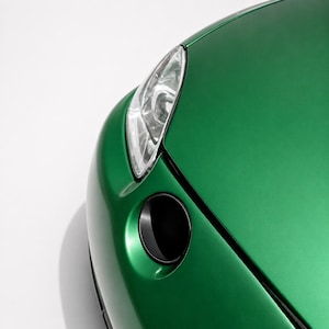 May include: A close-up view of the front corner of a metallic emerald green car, showcasing the smooth, curved hood, a clear headlight assembly with internal lens details, and a circular black air intake vent integrated into the body panel.