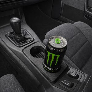 May include: A black Monster Energy drink can with a bright green claw logo and the text TAURINE, positioned in the center console of a vehicle next to a black gear shift knob and two rectangular power window switches on a textured black surface.