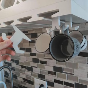 May include: A white plastic under-cabinet mug hook designed to slide onto a shelf edge. The hook features a T-shaped profile with a curved end for hanging mugs. Several similar white hooks are installed on the cabinet, holding ceramic mugs.