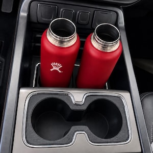 May include: Two red metal water bottles with a white logo featuring a person jumping over a wave, placed inside the cup holders of a vehicle center console. The console features a black plastic finish with chrome trim and various control buttons.
