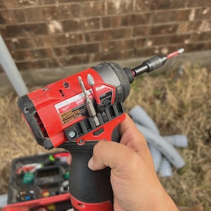 May include: A red and black cordless impact driver featuring a black rubberized grip, a metal chuck holding a drill bit, and a storage rail on the side containing two additional bits. The tool body displays white text and warning labels.