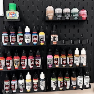May include: A black pegboard wall organizer holding various hobby paint bottles and jars. The collection includes Vallejo Game Color, Model Color, and Game Air paints, along with Citadel Colour pots and a Tamiya Extra Thin Cement bottle.