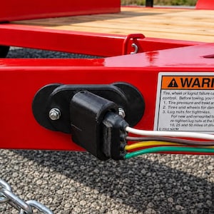 May include: A bright red utility trailer frame featuring a black plastic electrical connector plug with white, brown, yellow, and green wires attached. A rectangular warning label with black text is affixed to the side of the red metal frame.