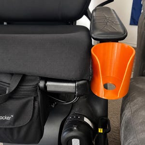 May include: A bright orange cylindrical cup holder attached to the side of a black power wheelchair. The wheelchair features a black padded seat, a black armrest with a textured surface, and a black storage bag with the partial text acker visible.