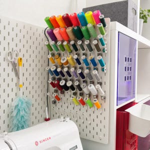 May include: A white pegboard wall organizer holding numerous colorful sewing thread spools in various shades including red, blue, green, yellow, and pink. A pair of scissors and a light blue duster hang on the side, above a white Singer sewing machine.
