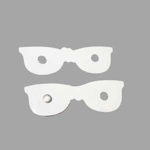 Puede incluir: Dos recortes blancos y planos con forma de gafas, hechos de un material liso. Cada recorte presenta dos aberturas circulares donde estar&iacute;an los cristales. Un recorte tiene un peque&ntilde;o im&aacute;n met&aacute;lico circular adherido a la superficie frontal.