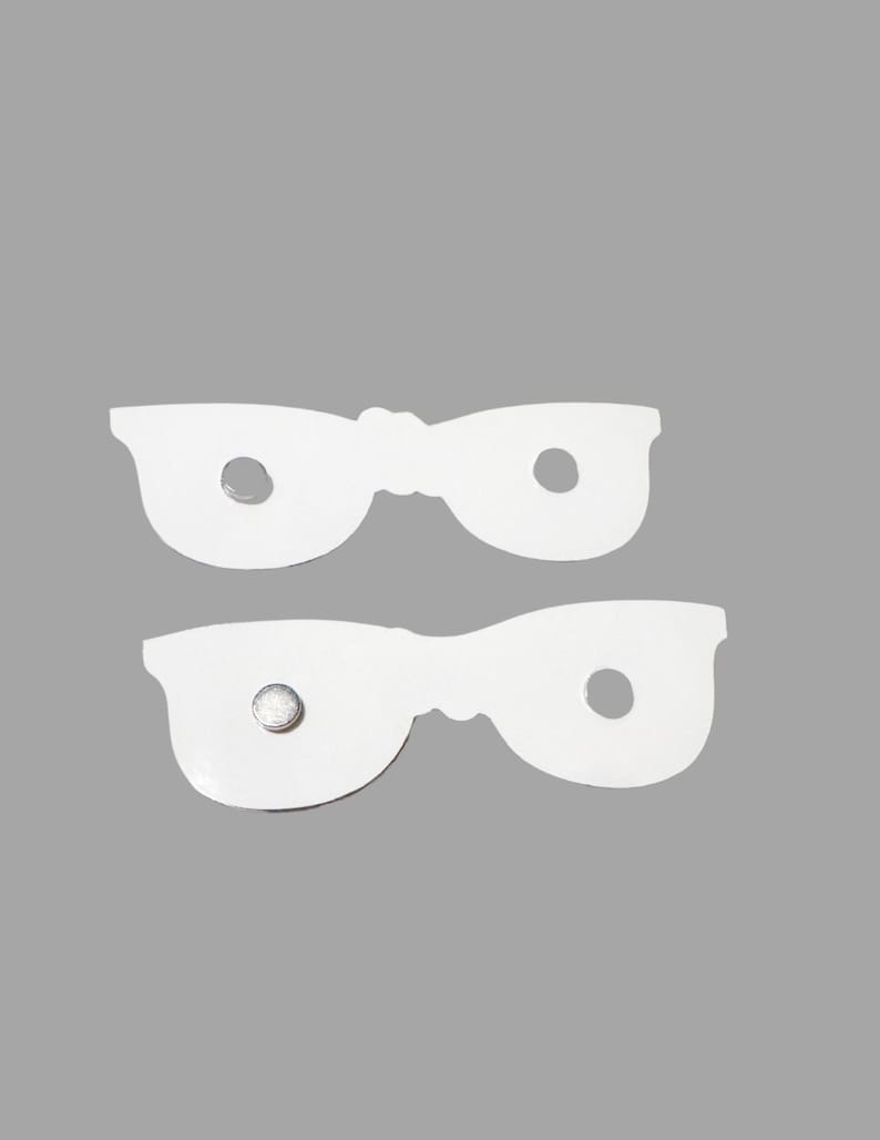 Puede incluir: Dos recortes blancos y planos con forma de gafas, hechos de un material liso. Cada recorte presenta dos aberturas circulares donde estar&iacute;an los cristales. Un recorte tiene un peque&ntilde;o im&aacute;n met&aacute;lico circular adherido a la superficie frontal.