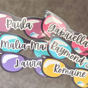 Puede incluir: Una colecci&oacute;n de pegatinas troqueladas con forma de gafas de sol, cada una con un nombre &uacute;nico en letra blanca: Paula, Gabriella, Malia-Marie, Raymond, Laura y Romaine. Las lentes muestran colores vibrantes, iconos de cruceros y palmeras.