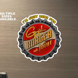 May include: A circular sticker resembling a bottle cap, featuring the text "I'M JUST HERE FOR" at the top. The center displays "Guy's! BURGER Joint" in gold lettering, with "EST 1968" below. The sticker is red, gold, and gray.