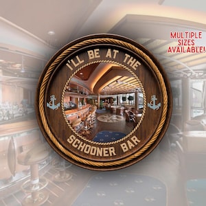 以下が含まれることがあります： ロープの縁取りと「I'LL BE AT THE SCHOONER BAR」という文字が特徴の円形木製看板。中央には、スツールやテーブルが置かれたバーの内部写真が表示されています。両サイドには水色の錨のアイコンが配置されています。