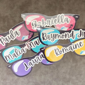 Puede incluir: Una colecci&oacute;n de gafas de sol de papel troquelado con varios nombres, incluidos Gabriella, Paula, Raymond Jr, Malia-Mari, Laura y Romaine. Cada par muestra un dise&ntilde;o de lente colorido con una ilustraci&oacute;n gr&aacute;fica de un crucero y palmeras.