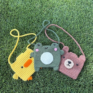 May include: Three handmade crochet bags in the shape of a yellow chick, a green frog, and a pink bear. Each bag has a long strap and is set against a green grass background. The bags have embroidered details.