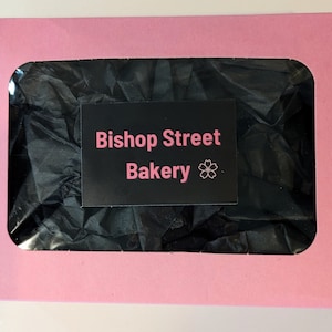 A pink pastry box with a clear cellophane window that shows black tissue paper inside the box. On top of this clear cellophane window sits a black sticker that says Bishop Street Bakery.