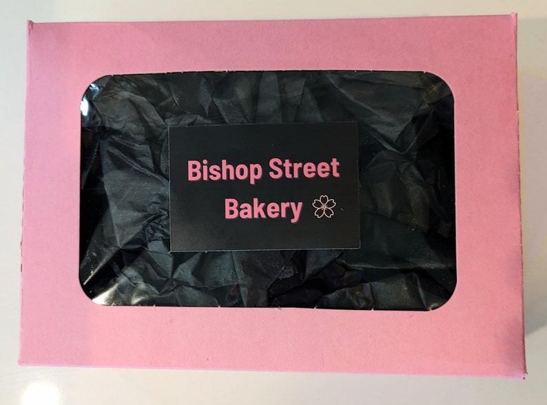 A pink pastry box with a clear cellophane window that shows black tissue paper inside the box. On top of this clear cellophane window sits a black sticker that says Bishop Street Bakery.