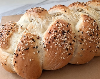 May include: Braided loaf of bread, golden brown with a variety of seeds sprinkled on top. The bread has a light, airy texture and is baked to a perfect golden color. The seeds include white sesame, black sesame, and other spices.