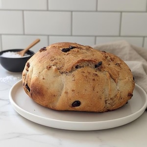 May include: A loaf of freshly baked bread with dark raisins, resting on a white plate. The bread has a golden-brown crust and a rustic appearance. A small black bowl of salt and a wooden spoon are in the background.
