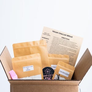 May include: A cardboard box filled with brown paper bags containing ingredients for making vegan challah bread. The ingredients include whole wheat flour, salt, turmeric, extra flour, and egg replacer. The box also includes a pink silicone pastry brush and a sticker with the text "Shop Street Bakery".